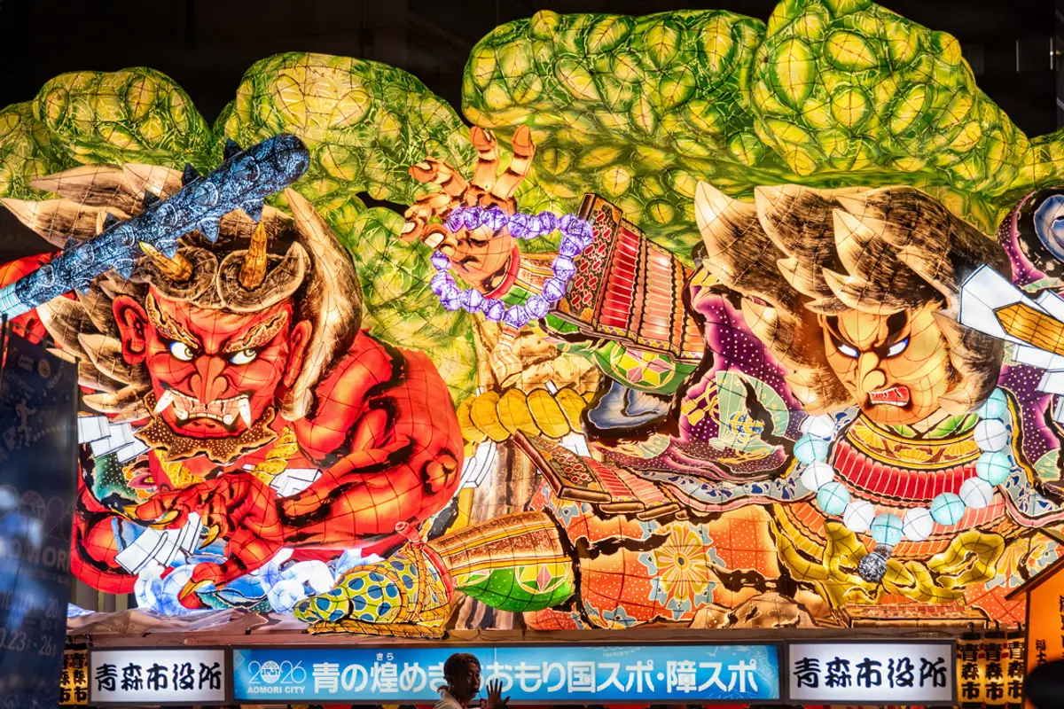 A huge illuminated Nebuta float on the night parade route in Aomori City, showing a red demon and an armoured warrior lit from the inside