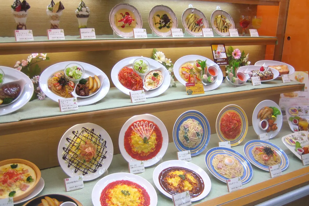 A restaurant display case filled with plastic models of pasta, rice dishes and parfaits, each beside a small price card in Japanese