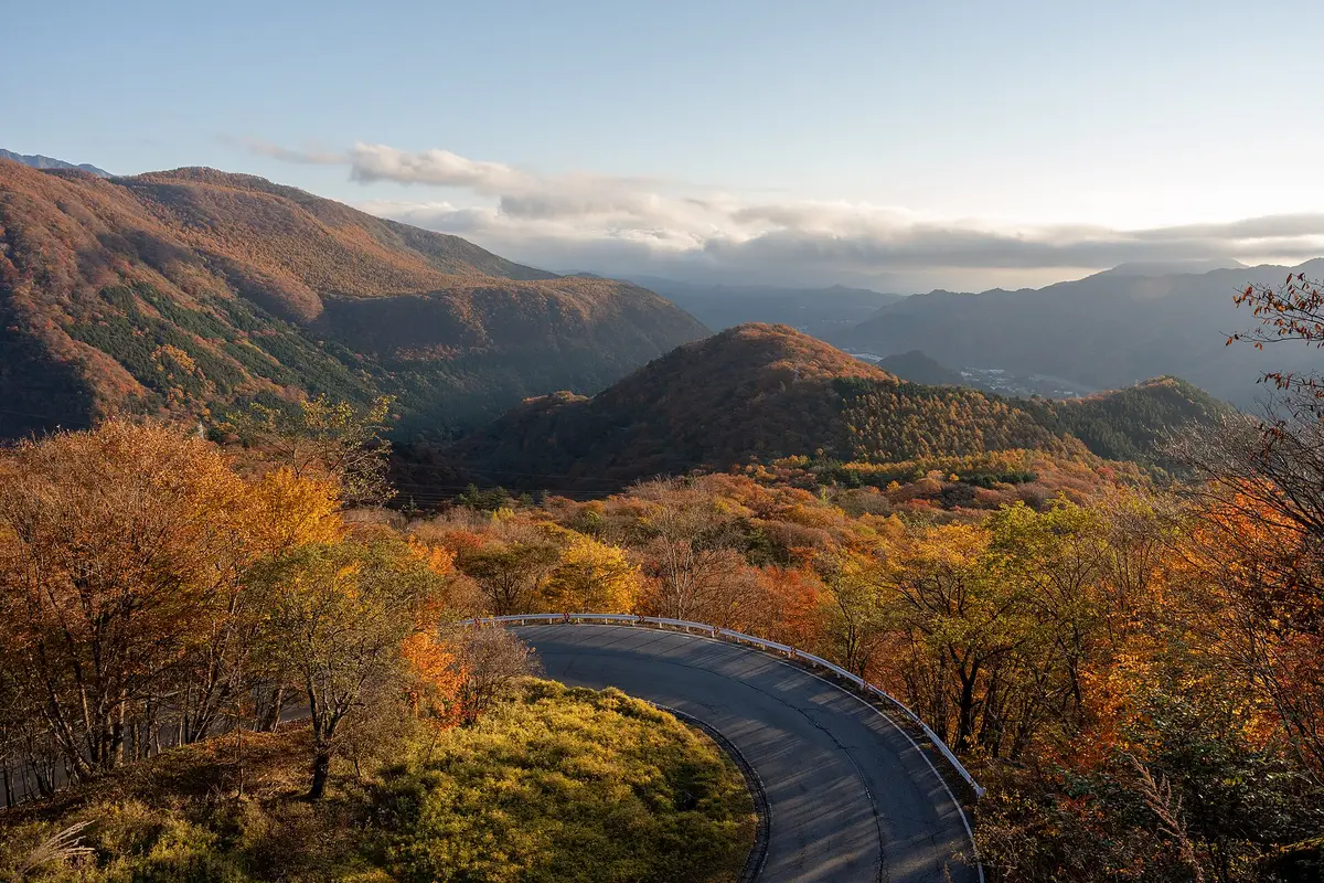 Looking down on a hairpin curve of the Irohazaka road as it winds through autumn forest, with mountain ridges beyond