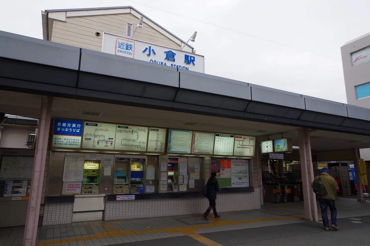 The exterior of Kintetsu Ogura Station, with its Japanese and English signage, ticket machines, and covered platform entrance