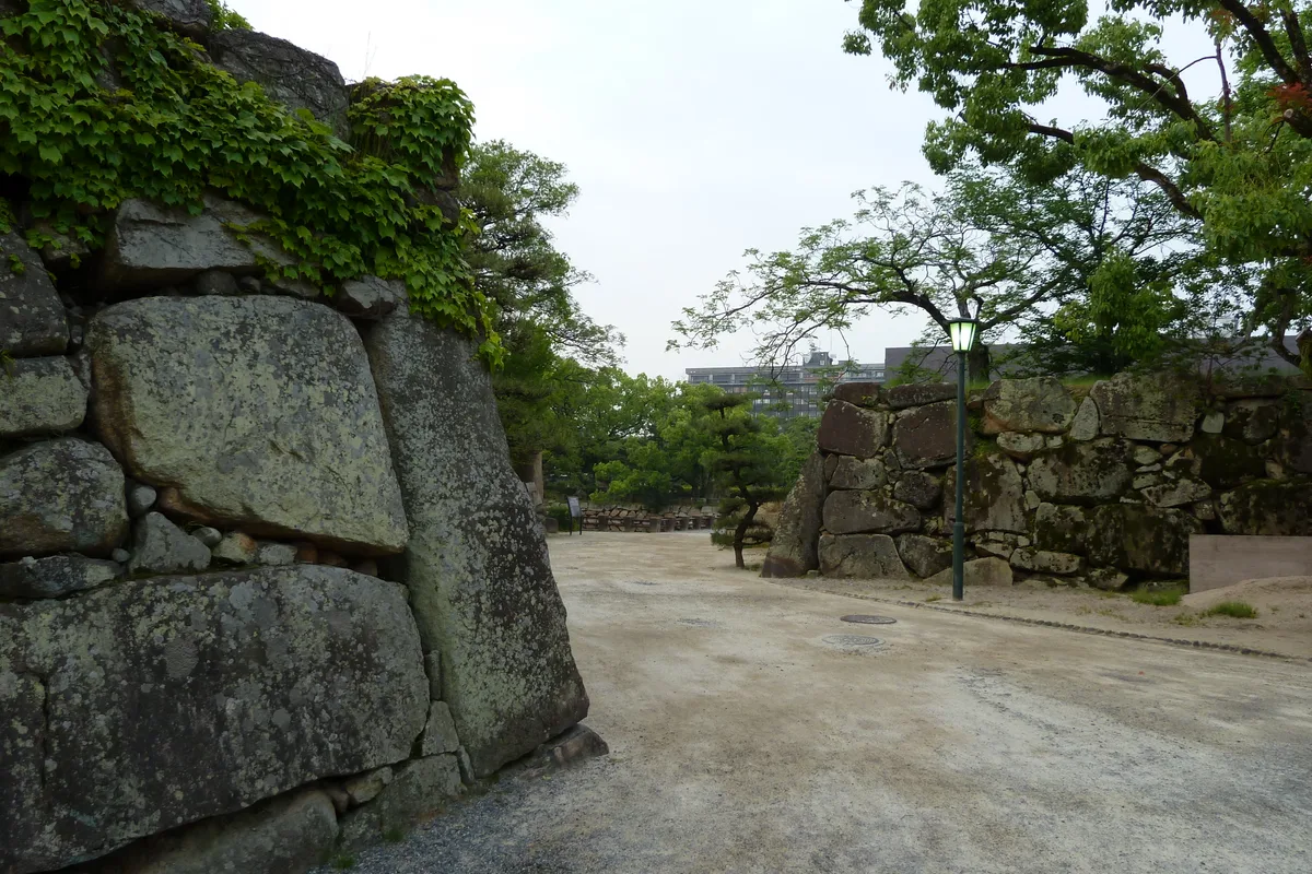 Large ivy-covered stone blocks of Okayama Castle's wall, with a second, visibly different stretch of stonework and a park lamp beyond