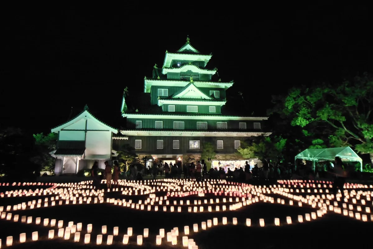 Okayama Castle's keep lit up green at night during the Ujo Togenkyo lantern festival, with rows of glowing lanterns covering the lawn