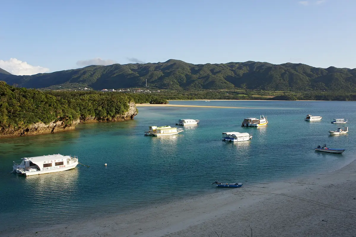 Seen from the hillside above Kabira Bay on Ishigaki Island: tour boats moored on turquoise shallows, jungle-covered ridges behind them, and white sand in the foreground