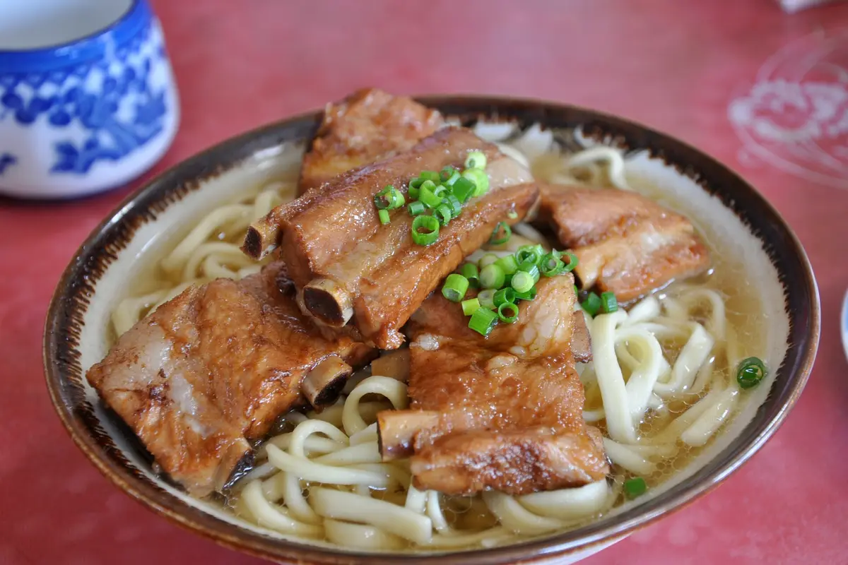 Close up on a bowl of Okinawa soba: thick wheat noodles in a clear broth, topped with stewed pork ribs and chopped green onion