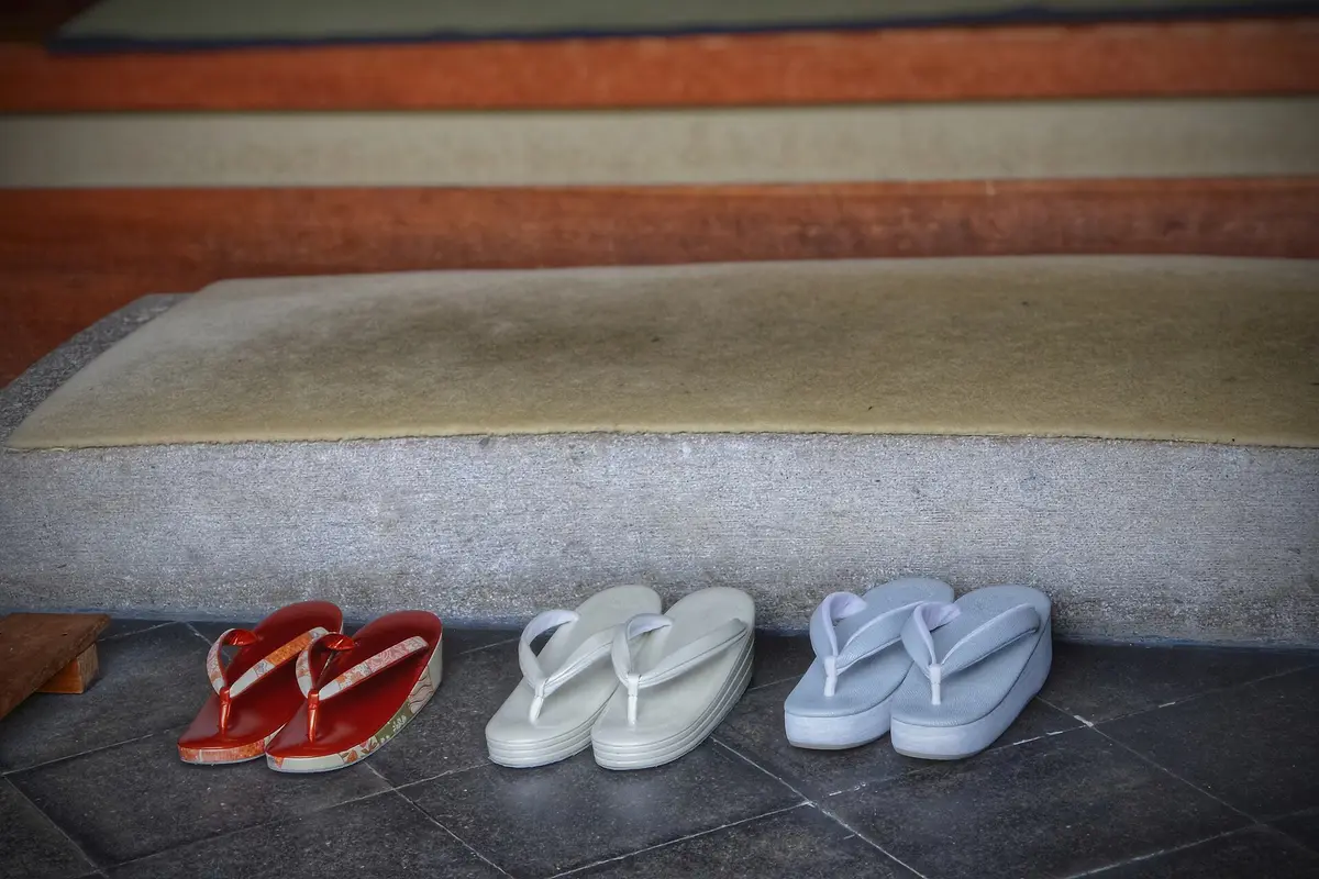 Three pairs of Japanese sandals set side by side on the stone floor of an entrance, below a raised step topped with a straw mat