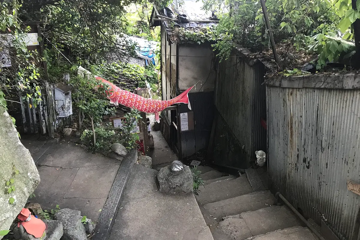 A narrow stone stairway on the Onomichi hillside, a carp streamer strung overhead, a painted cat stone beside the steps and corrugated fences on both sides