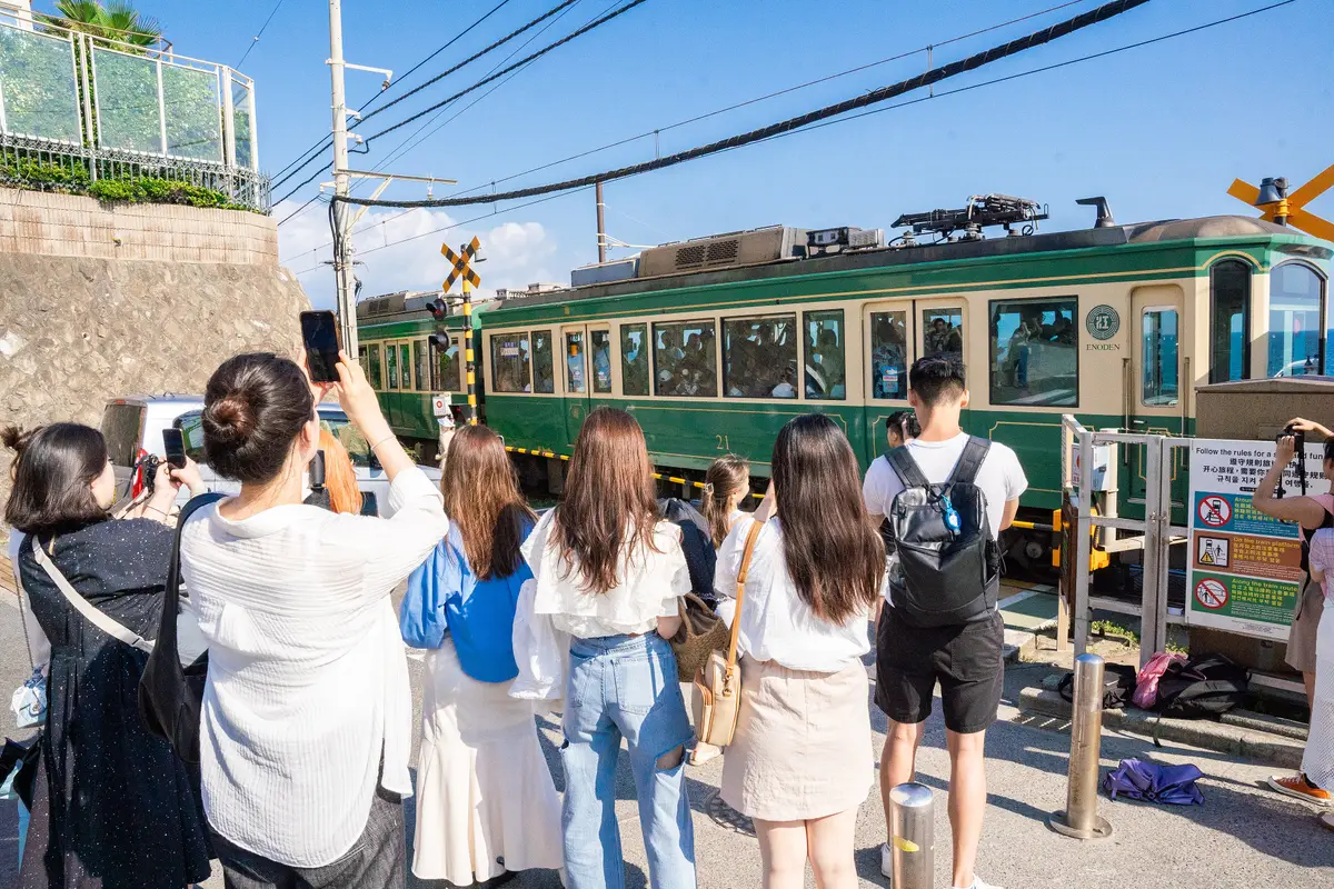Eine Reihe von Besuchern von hinten, die Handys hochhalten, während ein grüner Zug den Bahnübergang am Meer bei Kamakura-Kōkō-Mae passiert, daneben ein mehrsprachiges Schild