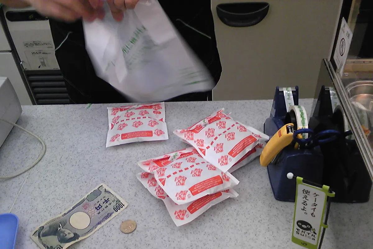 Hands opening a plastic bag over a convenience store counter, with several wrapped packets, a banknote and a coin laid out on the counter