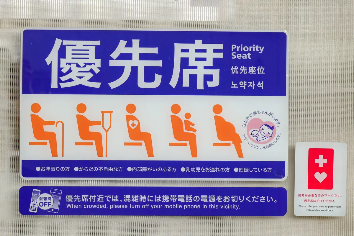 Close-up of a priority seat sign inside a Japanese train car showing five orange pictograms, with a separate white placard beside it displaying the red Help Mark