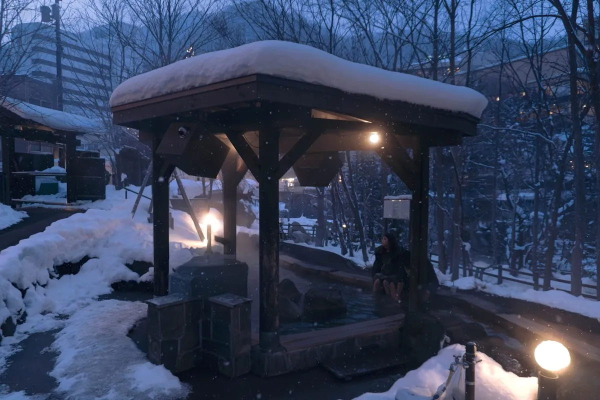 A snow-covered outdoor hot spring bath with warm lantern light on a winter evening in a Japanese onsen town