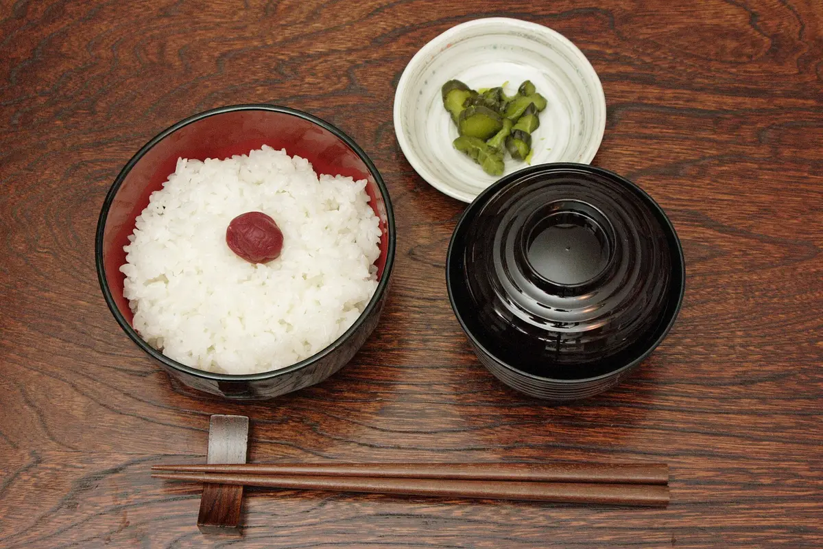 A bowl of white rice topped with a pickled plum, a covered soup bowl and a small dish of pickles on a wooden table, with a pair of wooden chopsticks laid flat on a wooden chopstick rest