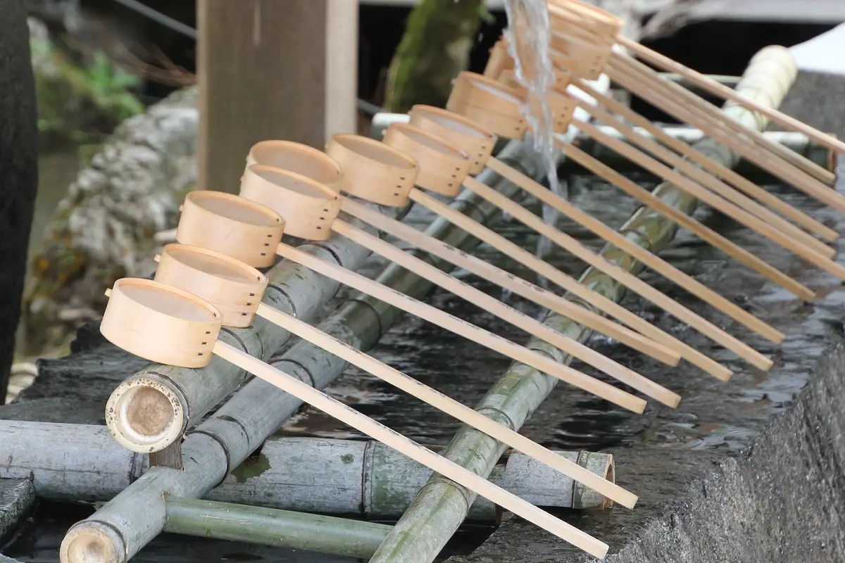 A row of wooden ladles laid across the bamboo rail of a stone purification basin at a Japanese shrine, with water running into the basin