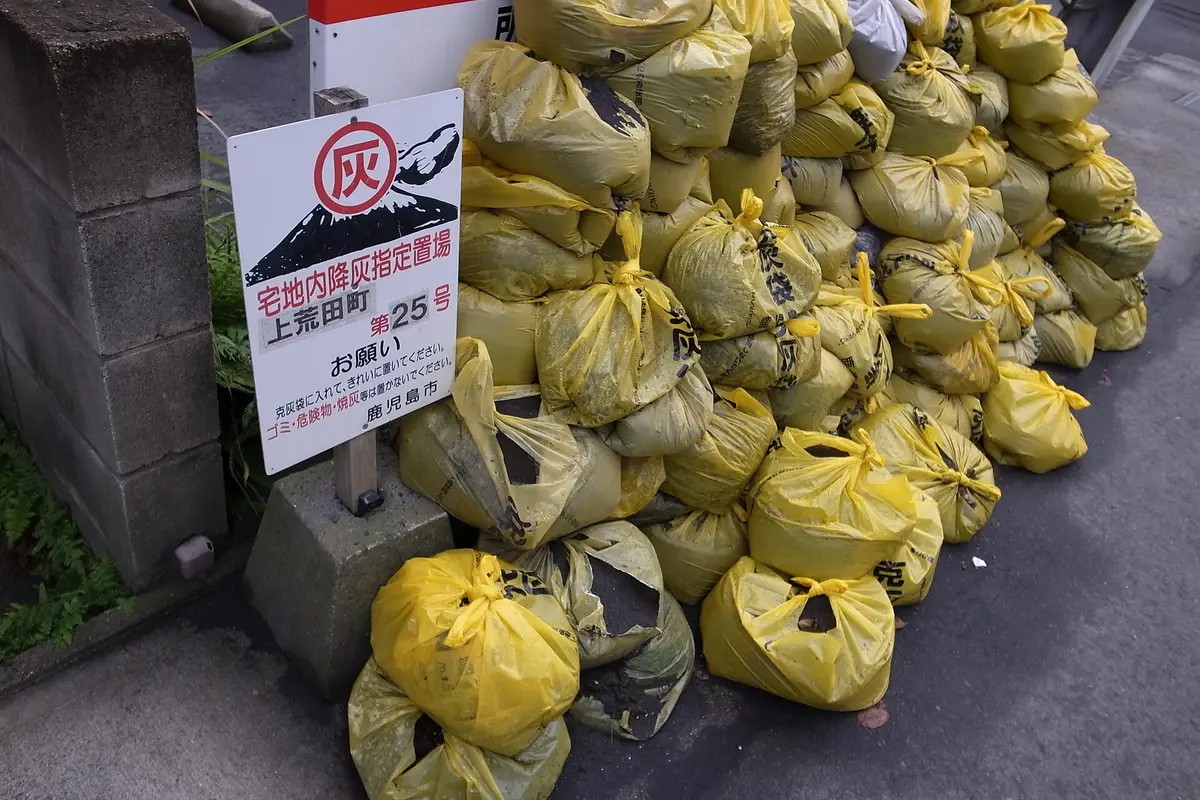Sacchetti gialli di cenere accatastati in un punto di raccolta designato lungo una strada di Kagoshima, accanto a un cartello comunale con un vulcano in eruzione