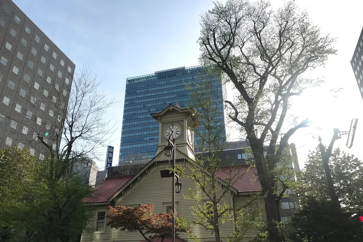 從街道高度仰望木造的札幌時計台，正後方矗立著一棟玻璃帷幕辦公大樓。