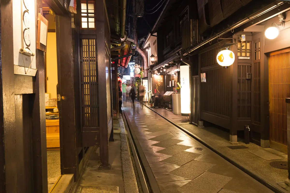 A narrow stone-paved lane in Kyoto at night, lined with wooden buildings, with blurred figures walking away in the distance