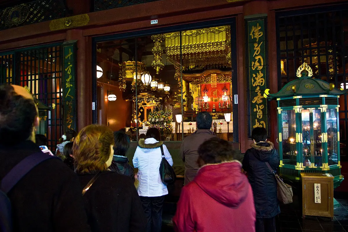 Visitors seen from behind, standing inside a temple hall in Tokyo and facing the lit altar