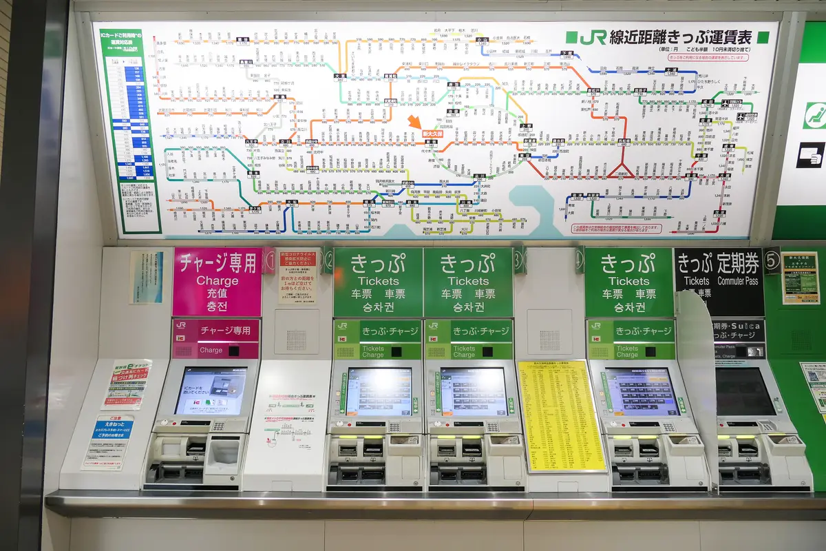 A large JR fare chart densely printed with station names and prices, mounted above a row of ticket machines at a Tokyo station