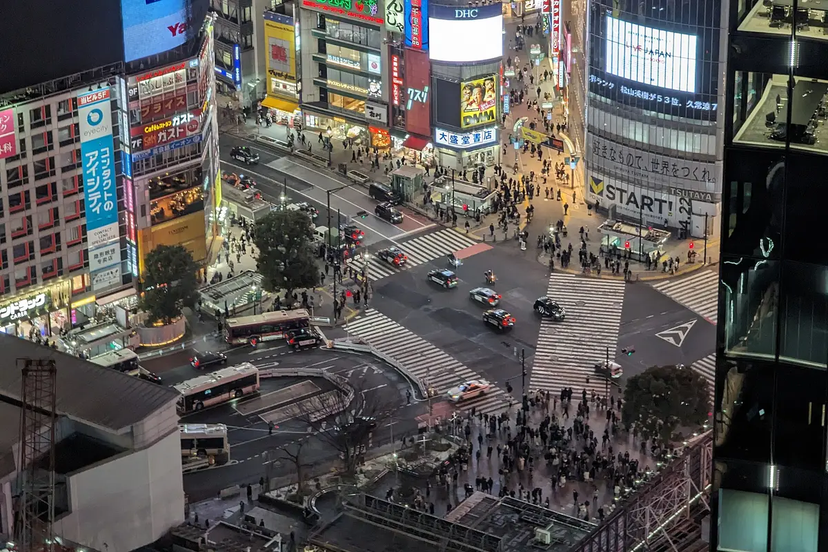 Seen from a nearby building after dark: the Shibuya Scramble Crossing with its big screens lit and crowds gathered on every corner