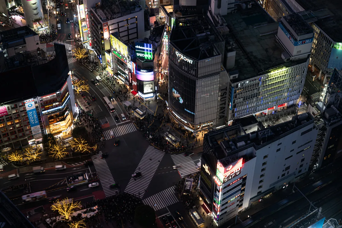 Night-time aerial view straight down onto the illuminated Shibuya scramble crossing, looking like a miniature model from above