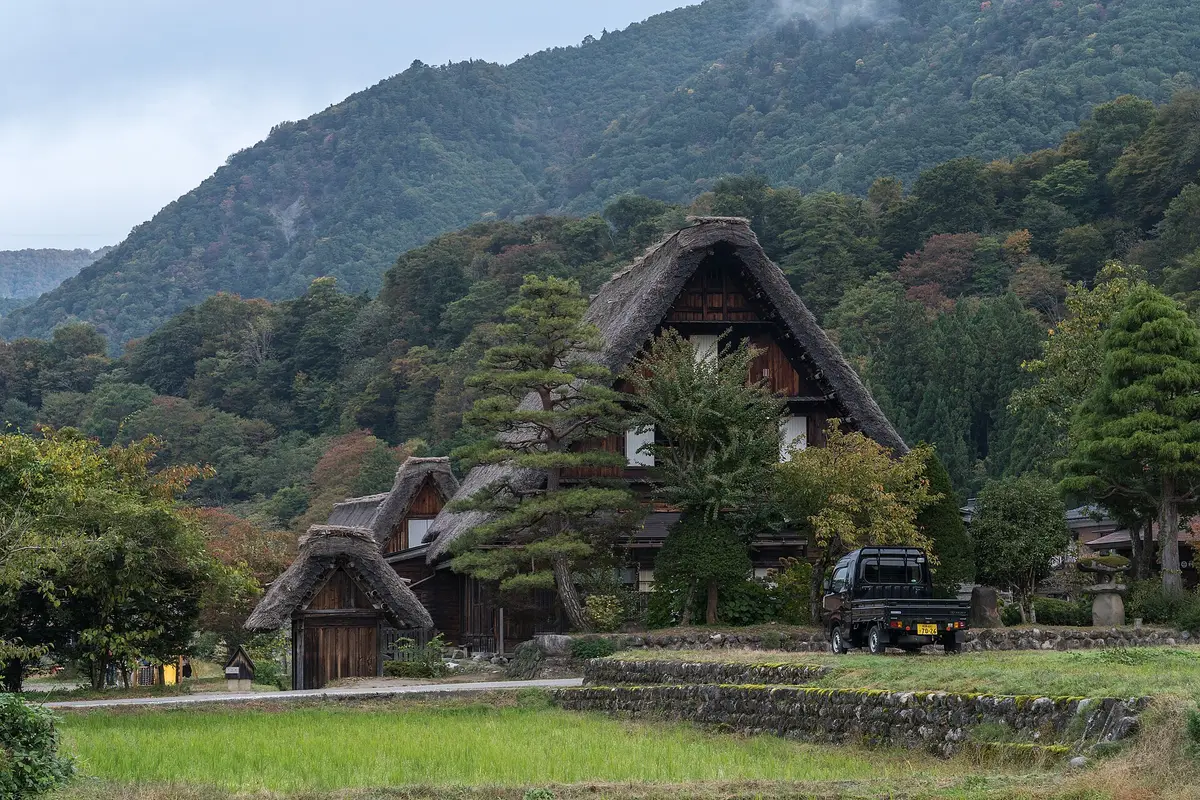 From the edge of a field in autumn: a gassho-zukuri farmhouse in Ogimachi with a small truck parked beside it and wooded mountains rising behind