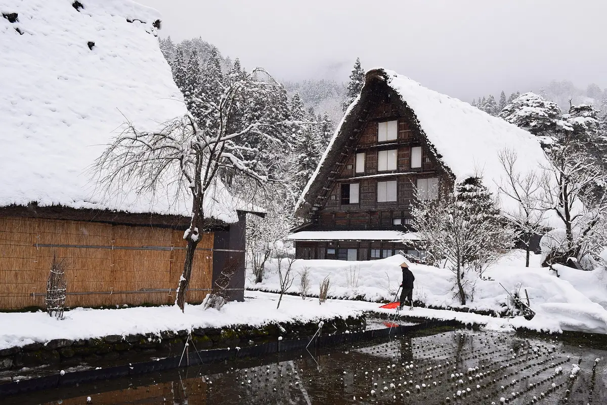 Deep snow at village level in Shirakawa-go: a gassho-zukuri farmhouse behind a straw snow screen, with one person clearing snow along the water's edge