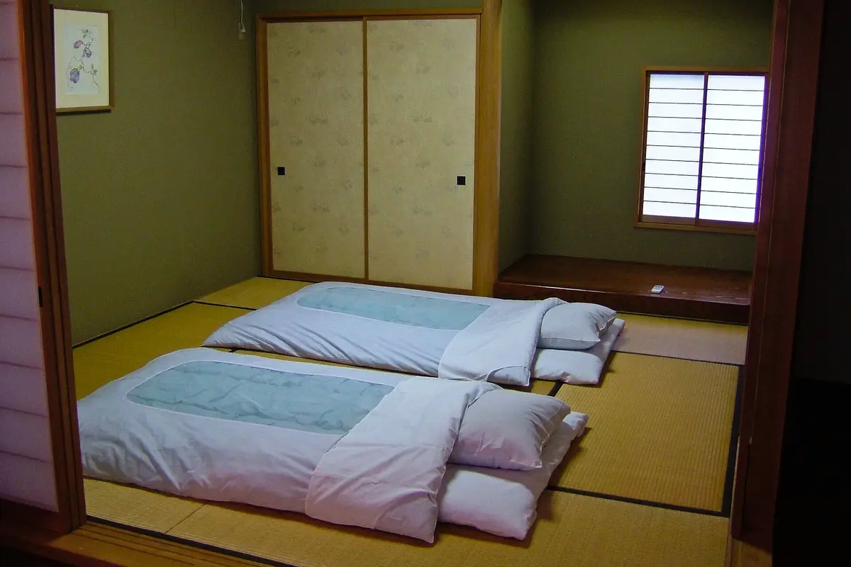 Two futons with quilts and pillows laid out directly on the tatami mat floor of a Japanese room, with sliding paper doors behind