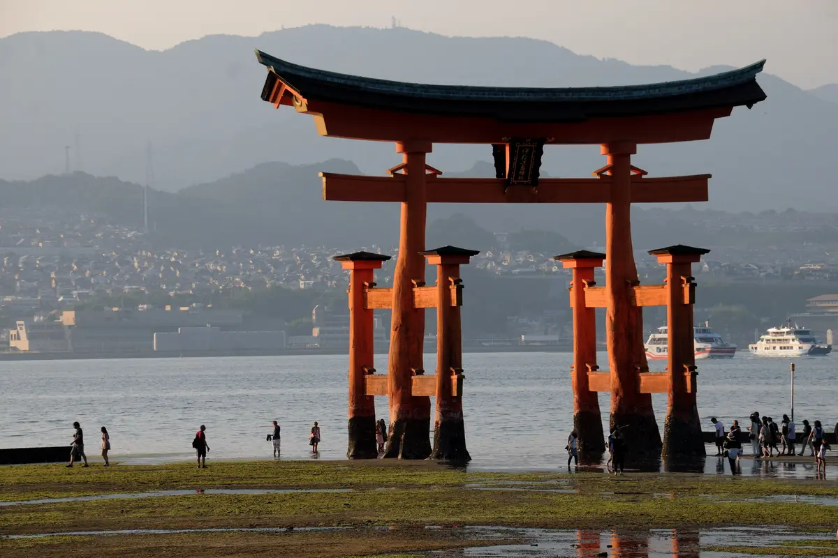 The great vermilion torii of Itsukushima Shrine at low tide, its pillars standing on exposed seabed with small figures walking nearby