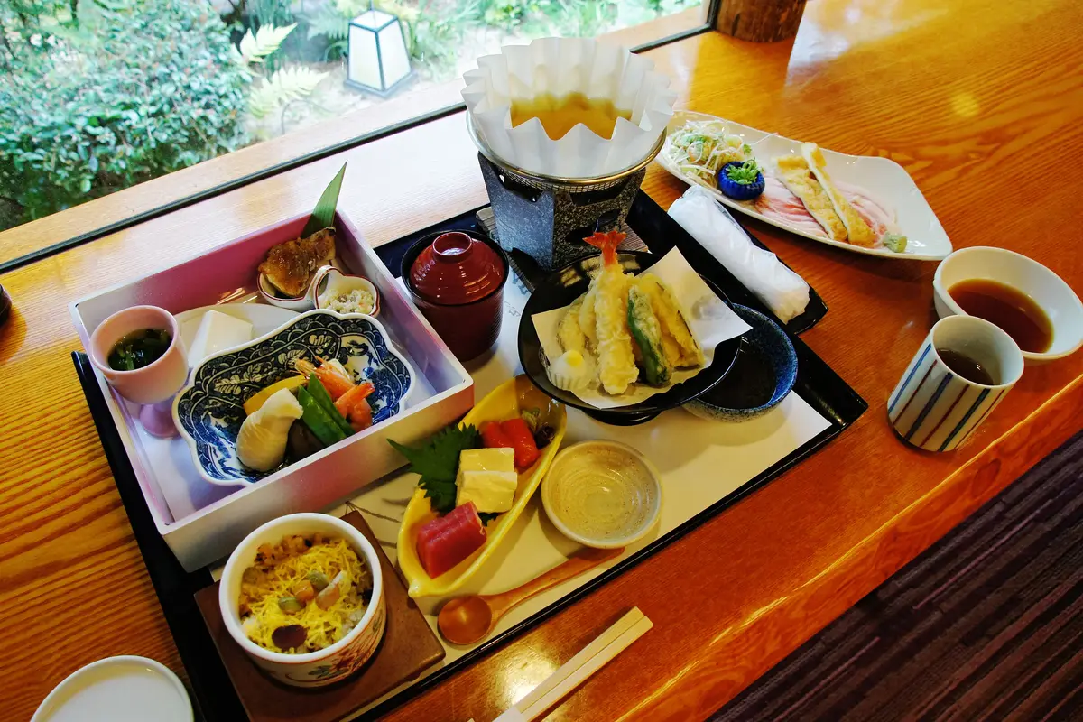 A formal Japanese meal set out on a black lacquer tray beside a garden window, with a tiered box of small dishes, a tempura plate, sashimi, rice and a lidded soup bowl