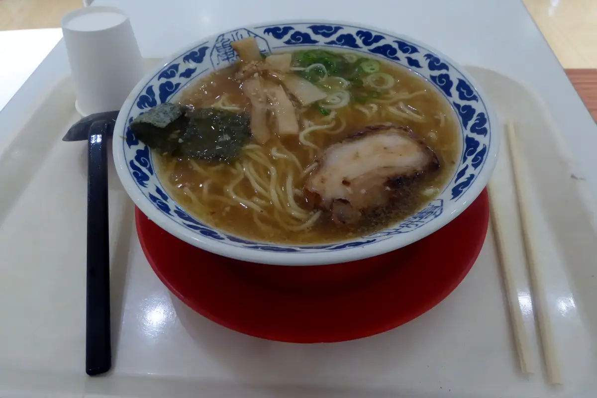 A bowl of ramen in a brown broth topped with sliced pork, bamboo shoots, spring onion and nori, on a white tray with a black spoon on the left and wooden chopsticks on the right