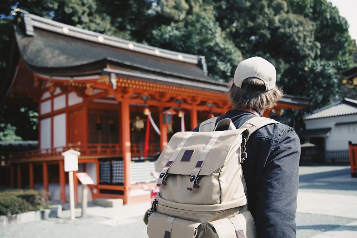 A solo traveler with a backpack gazing at a red shrine surrounded by trees in Japan