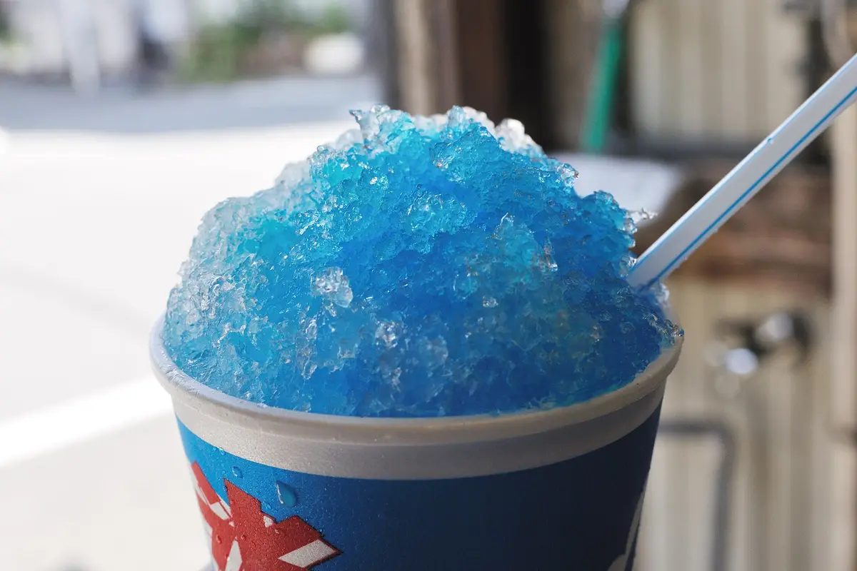 A paper cup heaped with shaved ice soaked in bright blue syrup, with a plastic spoon straw