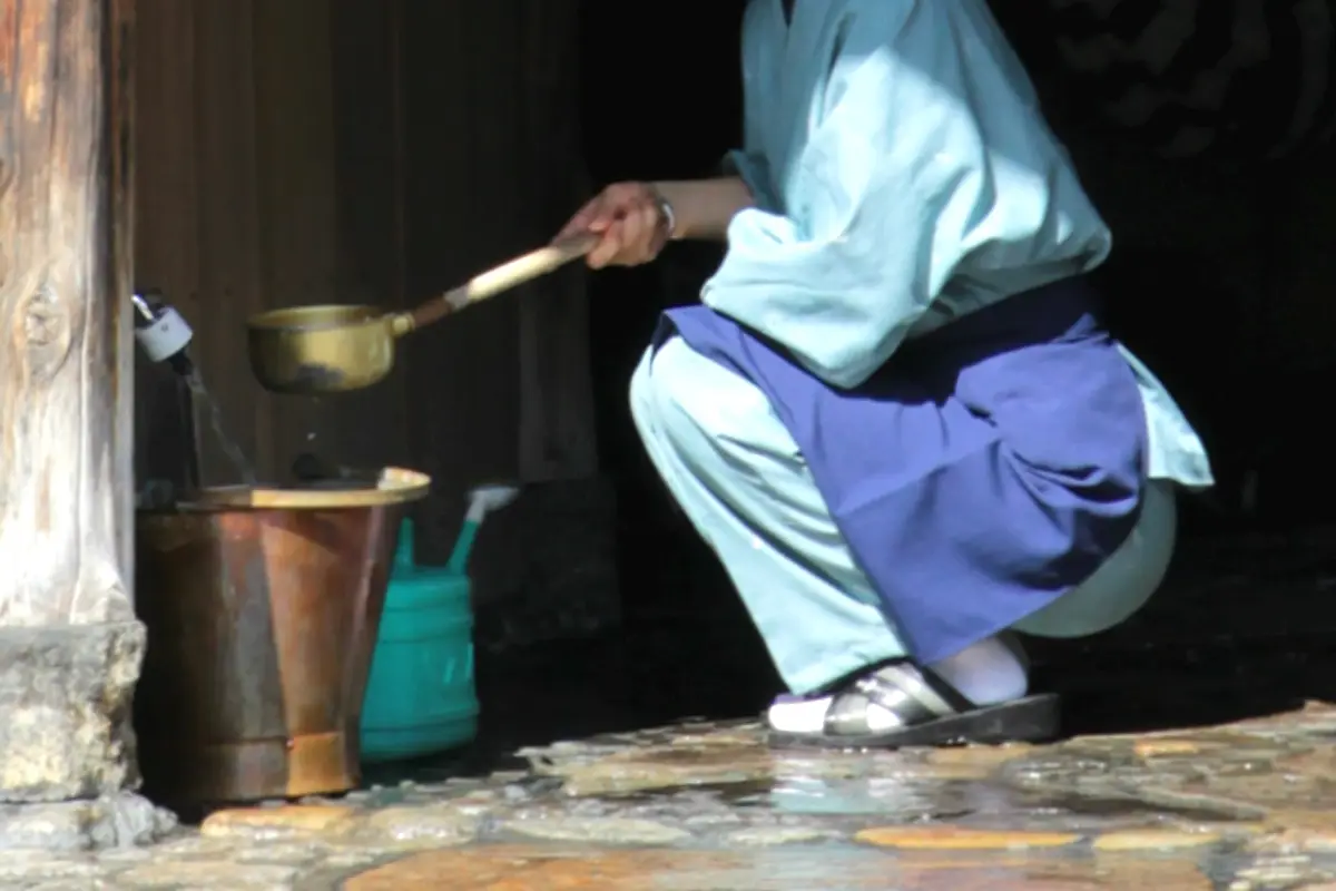 A long-handled metal ladle held over wet stone paving beside a copper bucket at a shop entrance