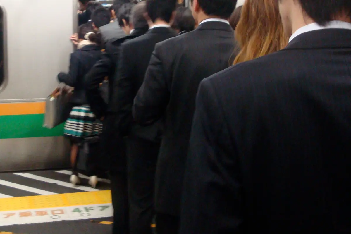 Commuters seen from behind, waiting along a platform and stepping one after another through the doorway of a full train