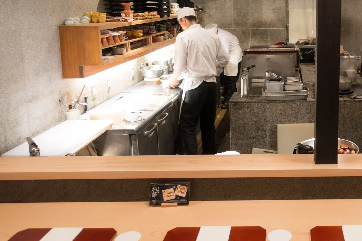 Two restaurant staff seen from behind at an open kitchen across a wooden counter, with place settings laid out in the foreground