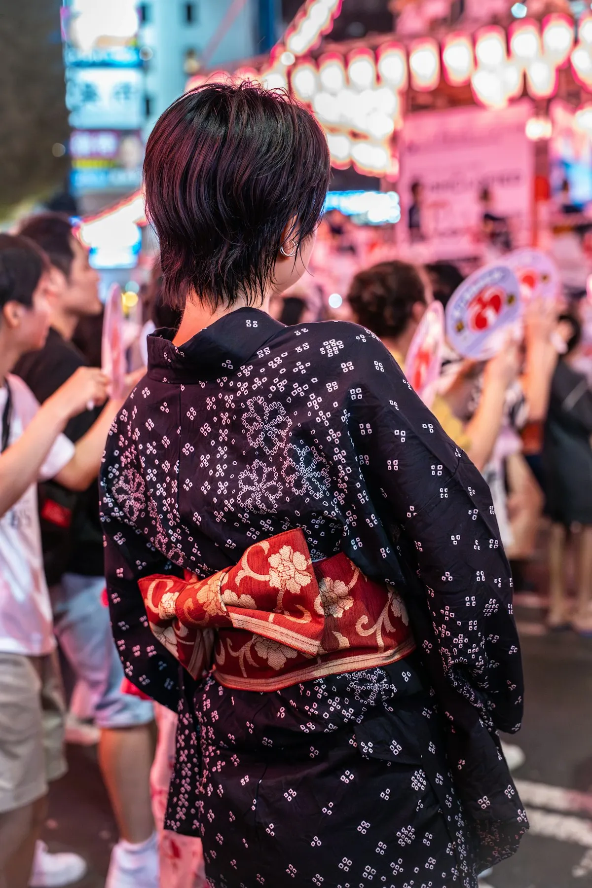 A woman in a dark yukata with a red obi seen from behind at a Japanese summer festival