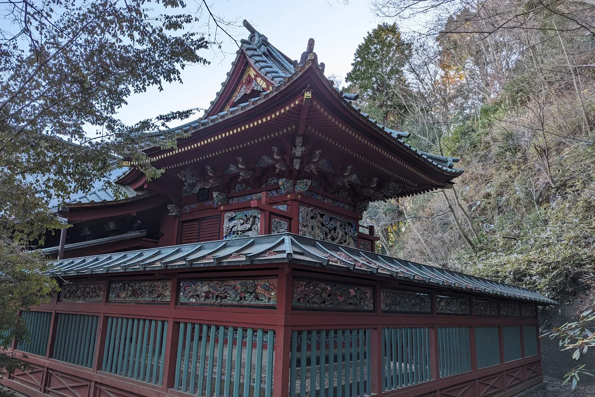 Eine kunstvolle, zinnoberrote Halle mit geschnitzten Drachenreliefs und blauem Ziegeldach, Teil des Yakuoin-Tempelkomplexes auf dem Takao-san, umgeben von kahlen Herbstbäumen