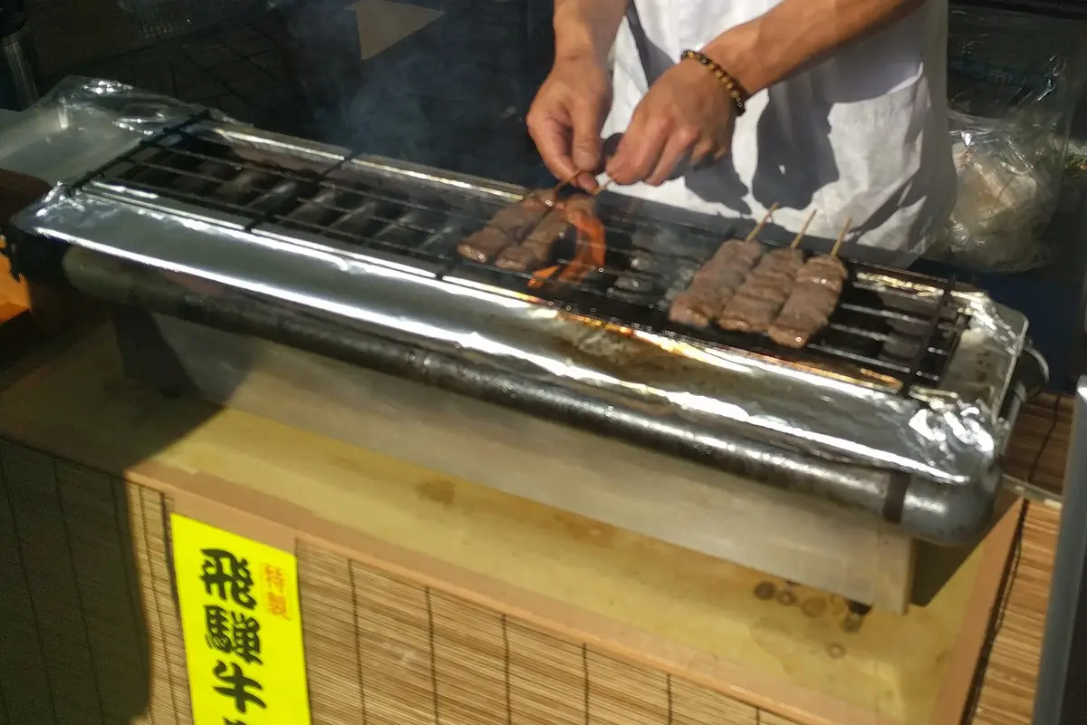 Hida beef skewers cooking on a small grill at a street stall in Takayama, with a vendor's hands turning one of them