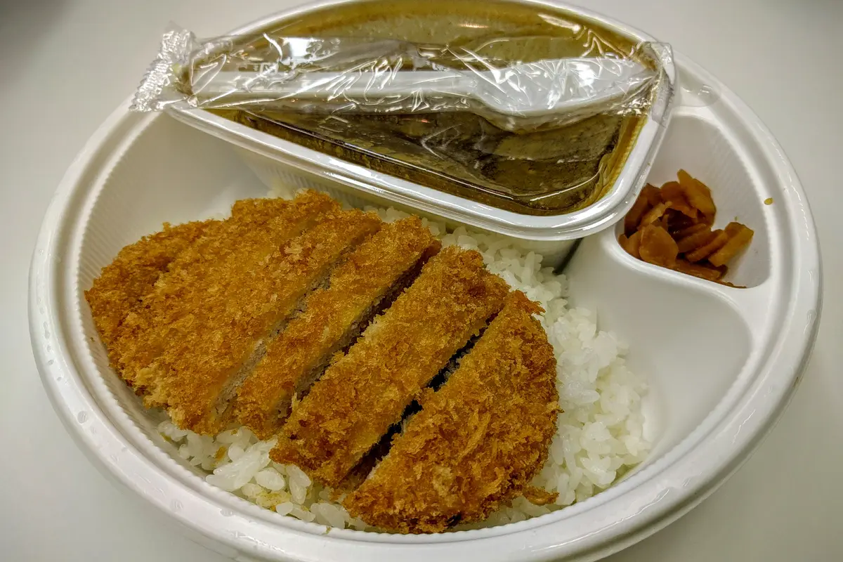 A breaded cutlet and rice in a partitioned disposable takeaway tray, with the sauce in its own lidded container and a wrapped spoon