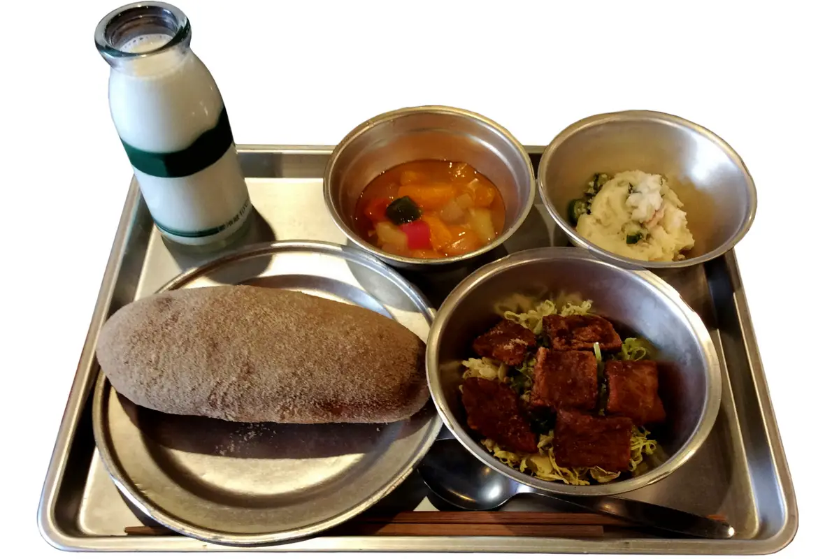 A reconstructed 1970s Japanese school lunch on a steel tray: a glass bottle of milk, a bread roll, a stewed dish, potato salad and a fried dish on shredded cabbage