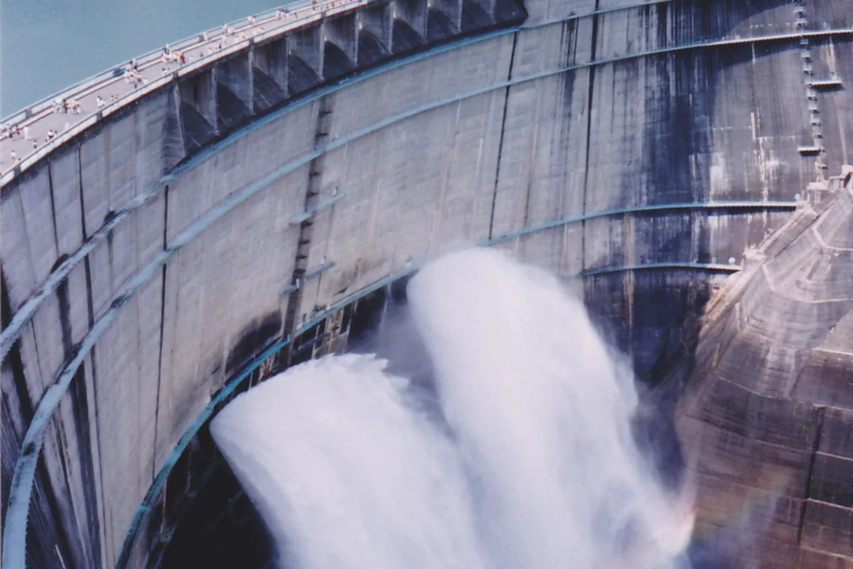 Der Kurobe-Staudamm lässt eine riesige Fontäne aus Sprühwasser ab, im Dunst ist ein schwacher Regenbogen zu sehen, oben auf der Staudammkrone gehen Besucher entlang