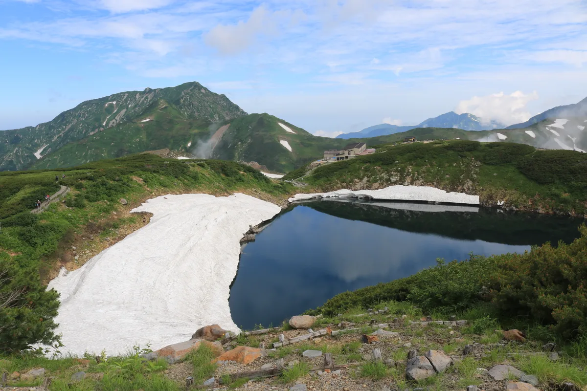 Mikuriga-ike, ein stiller, dunkler Bergsee mit einem Schneefleck, in dessen Oberfläche sich nahe Murodo grüne Sommerberge spiegeln