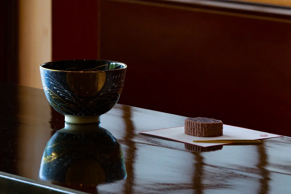 Eine dunkle Teeschale mit gemaltem Mond und Gräsern steht auf einem polierten Tisch, daneben eine kleine Süßigkeit auf gefaltetem Papier mit einem Holzstäbchen
