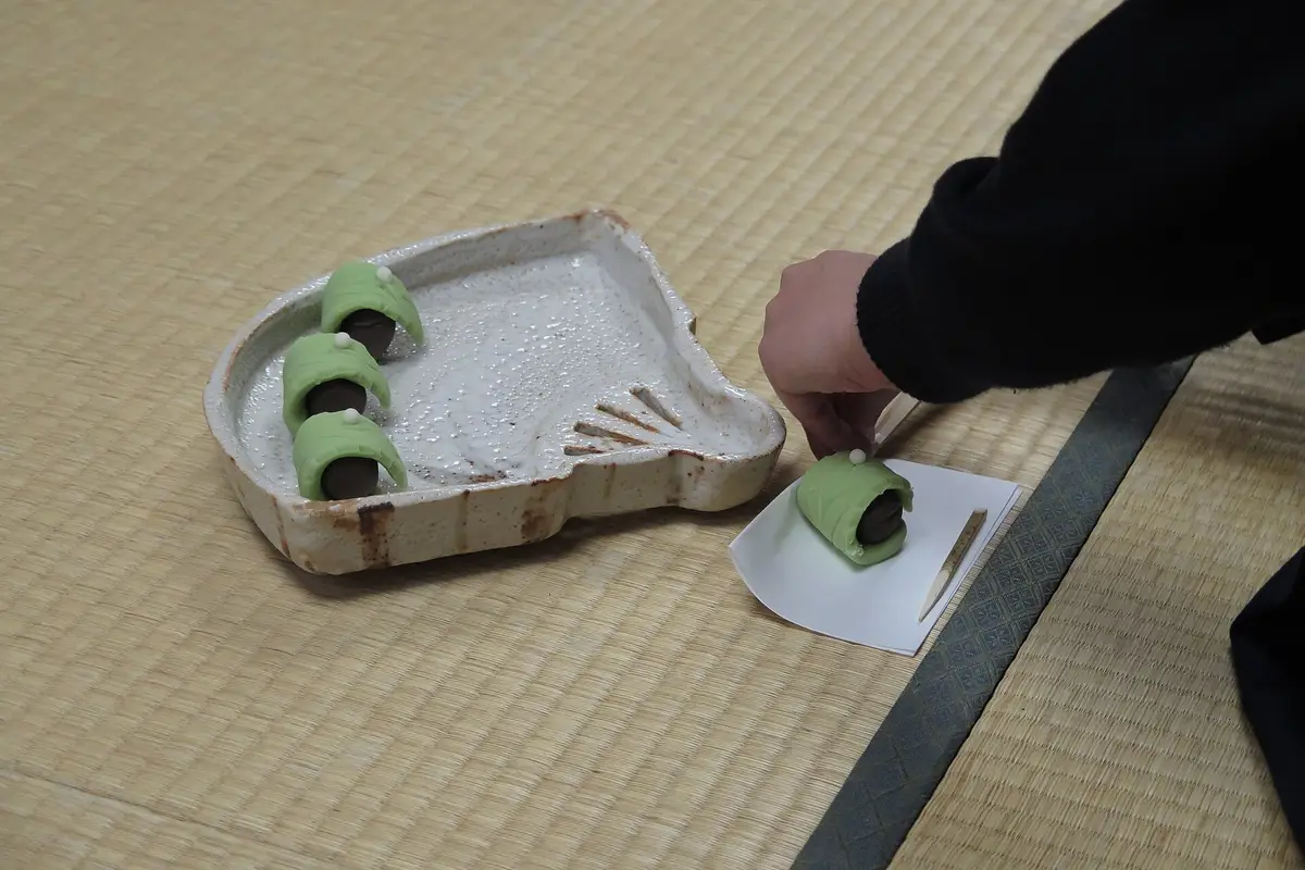 Auf dem Tatami hebt eine Hand ein Holzstäbchen von einer grünen Süßigkeit, die auf einem kleinen gefalteten Papier liegt, daneben ein Keramiktablett mit drei weiteren