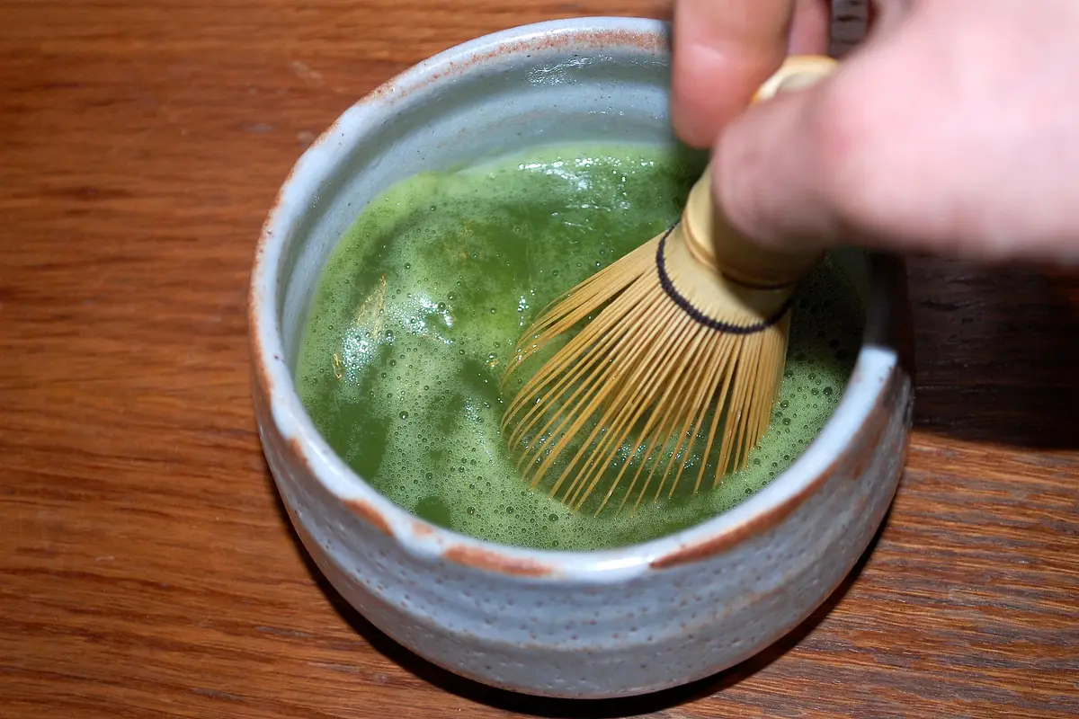 Von oben gesehen, eine Hand schlägt mit einem Bambusbesen leuchtend grünen Matcha in einer hellen Keramikschale auf