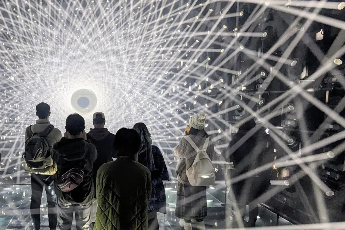 Visitantes de espaldas a la cámara dentro de una sala atravesada por líneas de luz en teamLab Borderless, en Azabudai Hills, Tokio