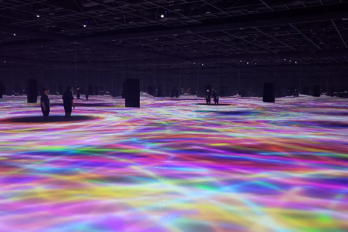Visto desde el otro extremo de la sala: luz de colores que fluye por todo el suelo de teamLab Planets, en Tokio, con unos pocos visitantes de pie muy separados entre sí
