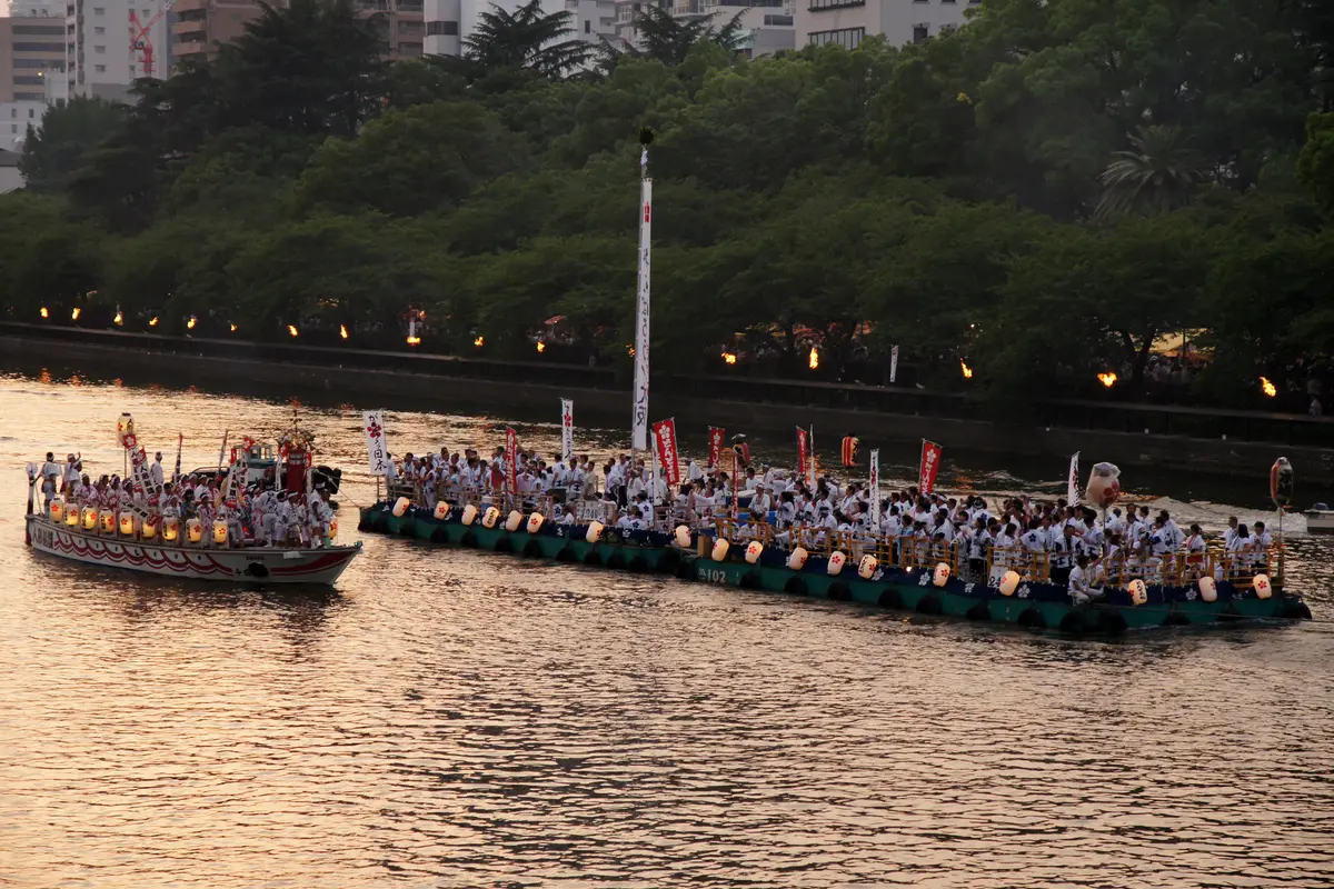 A lantern-hung boat under tow on the Ōkawa River, with a line of torches burning along the stone embankment