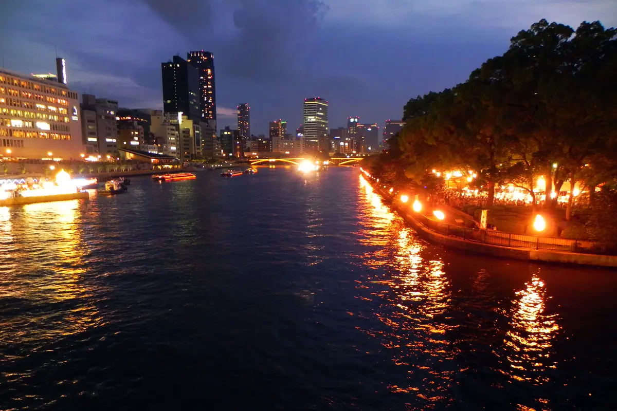 Dusk view from a bridge along the Ōkawa, with torches burning down the right bank and people gathered under the trees