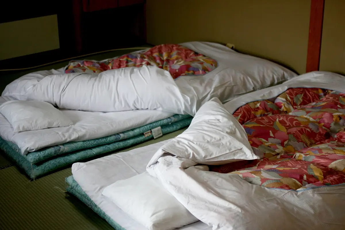 Auf dem Tatami-Boden eines japanischen Gasthauszimmers ausgelegte Futons mit weißen Kopfkissen und gemusterten Decken