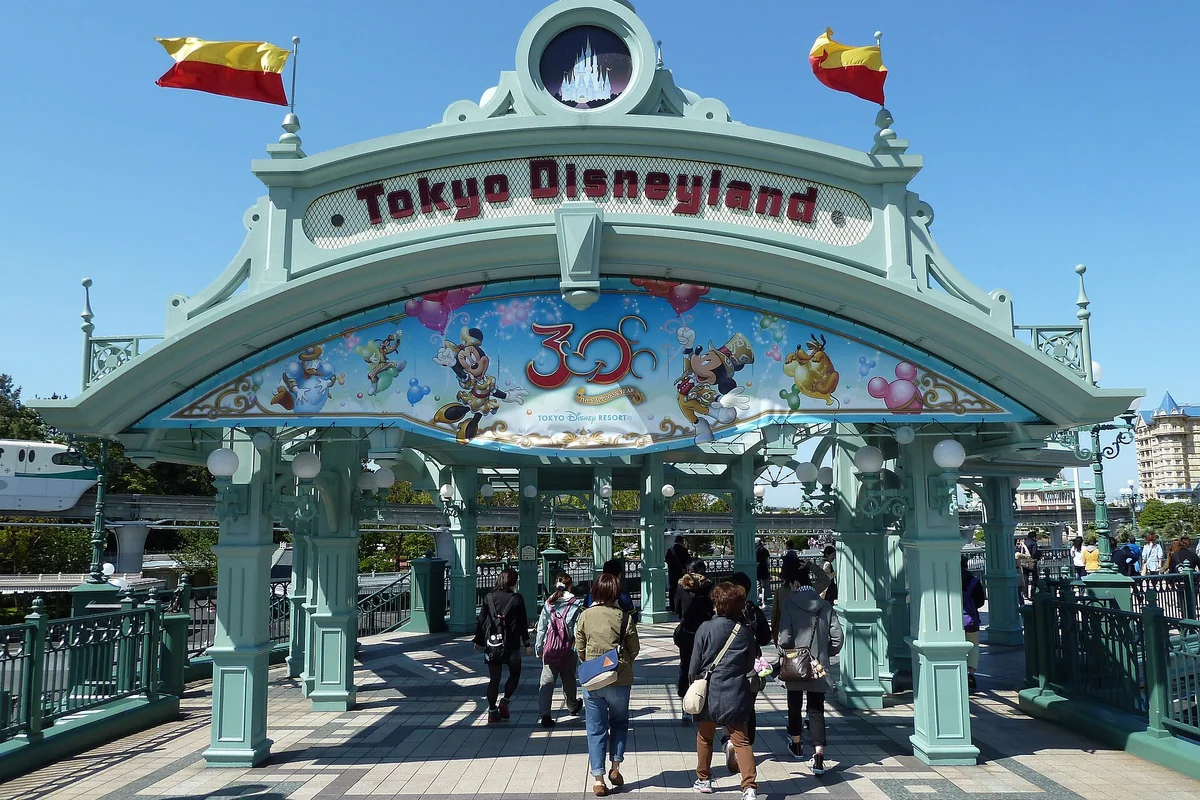 Visitors walking under the Tokyo Disneyland entrance gate archway with the park's monorail passing in the background